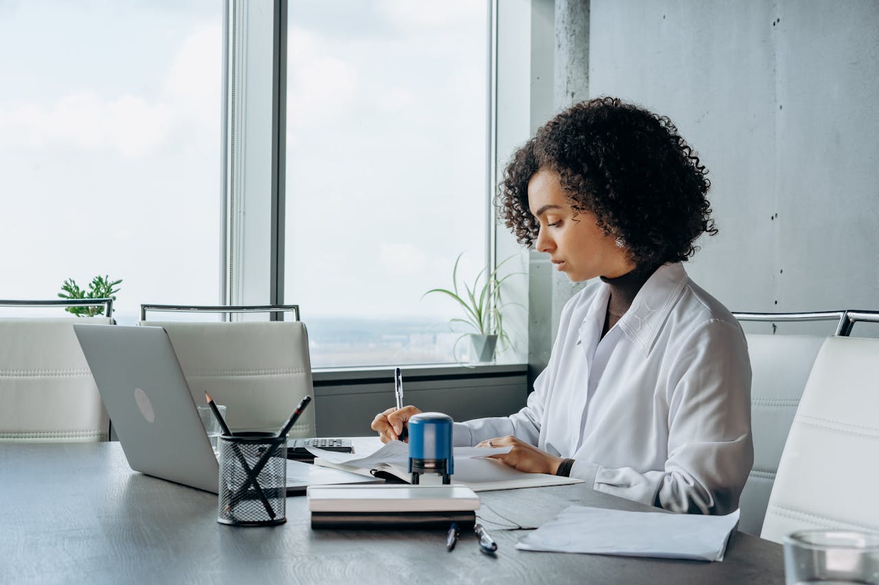African American woman in a white shirt working at a laptop in a bright, modern office setting.