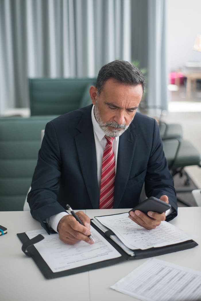 A professional businessman in a suit reads documents and checks his phone at a desk.